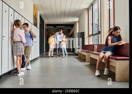 un gruppo di bambini sta nel corridoio accanto agli armadietti, mentre un insegnante li istruisce in un ambiente luminoso e vivace in classe. Foto Stock