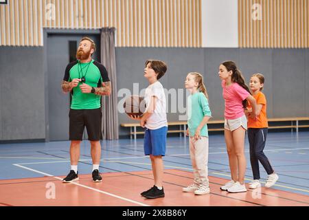 Un uomo si erge davanti a un gruppo di bambini, tenendo in mano un pallacanestro e fornendo indicazioni in un ambiente vivace e impegnato in classe. Foto Stock