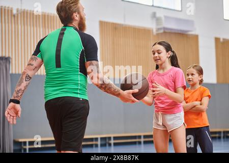 Un insegnante di sesso maschile si erge tenendo in piedi una pallacanestro davanti a un gruppo variegato di bambini in un ambiente luminoso e vivace in classe. Foto Stock