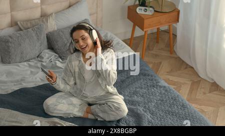 Vista dall'alto donna caucasica felice letto seduti cuffie smartphone telefono cellulare gadget tecnologia elettronica ascolto musica canto divertente energico Foto Stock