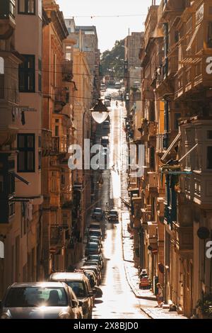 Le auto percorrono una strada ripida e stretta costeggiata da alti edifici storici a la Valletta, Malta Foto Stock