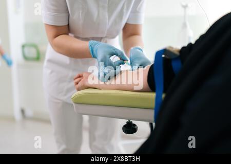 L'infermiere esegue l'infusione endovenosa alla paziente in clinica. Giovane medico in scrub inserti uniformi IV linea ago in vena di donna seduta Foto Stock