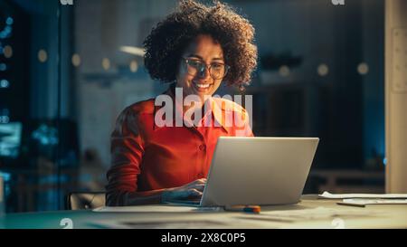 Giovane donna nera che lavora su un computer portatile in ufficio creativo la sera. Project Manager multietnico che scrive e-mail e si sorprende quando riceve un messaggio da un collega. Foto Stock