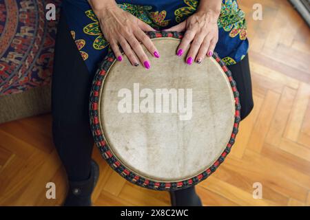 Vista dall'alto delle mani femminili sulla testa in pelle del tamburo djembe di una donna irriconoscibile seduta a giocare a casa mentre si pratica. con il pavimento sullo sfondo. Foto Stock