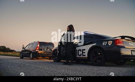 Agente di polizia di colore che esce dalla Patrol Car e si dirige verso una Pretted Over Car. I poliziotti rispondono a una chiamata 911 su un'auto sospetta bloccata sulla strada, indagando sulla situazione Foto Stock