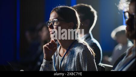 Primo piano Ritratto di giovane donna entusiasta che partecipa a un evento motivazionale, siede in una sala concerti affollata, si avvicina al palco e ascolta attentamente la presentazione. Foto Stock