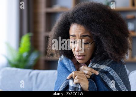 Una giovane donna è avvolta da una coperta blu a quadri, sentendosi fredda e mostrando una triste espressione. Ha gli occhiali e un taglio di capelli afro. Lo sfondo include una pianta verde e ripiani sfocati. Foto Stock