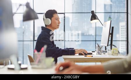 Giovane uomo asiatico che lavora su un computer e ascolta attraverso le cuffie in un ufficio moderno. Client Advisor maschio che risponde alle richieste. Grande finestra con vista sulla città sullo sfondo. Foto Stock