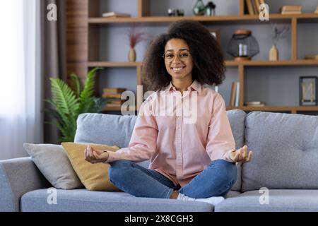 Una donna con capelli ricci e occhiali sta meditando su un divano grigio in un soggiorno con scaffale e decorazioni vegetali. Indossa una camicia rosa e jeans blu, seduta a gambe incrociate e sorridente. Foto Stock
