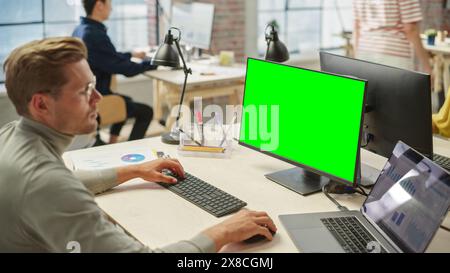 Ritratto di giovane uomo bianco che indossa gli occhiali che lavora su un laptop e un computer con schermo verde durante il giorno in un moderno Bright Office. Uomo d'affari che utilizza Chroma Key sul personal computer. Foto Stock