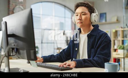 Ritratto di Asian Creative Young man focalizzato mentre utilizza il computer in Modern Office. Supervisore della logistica con cuffie accese, analisi di grafici, Borsa e dati finanziari Foto Stock