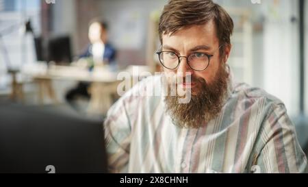 Ritratto di giovane uomo caucasico barbucato con gli occhiali concentrati e che lavora al computer in un ufficio luminoso e spazioso. Amministratore maschio sorridente dopo aver completato con successo un'attività. Foto Stock