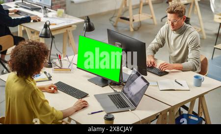 Gruppo diversificato di colleghi creativi che lavorano su computer in Modern Office. Donna birazziale che utilizza un Key Screen monitor Green Chroma. Designer di grafica maschile caucasica sorridente e digitante. Vista ad angolo alto. Foto Stock