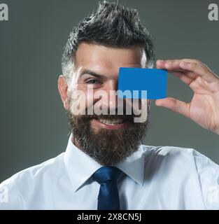Uomo sorridente con barba che copre l'occhio con un mockup vuoto biglietto da visita vuoto. Bell'uomo d'affari con carta di credito o di debito. Uomo barbuto felice dentro Foto Stock