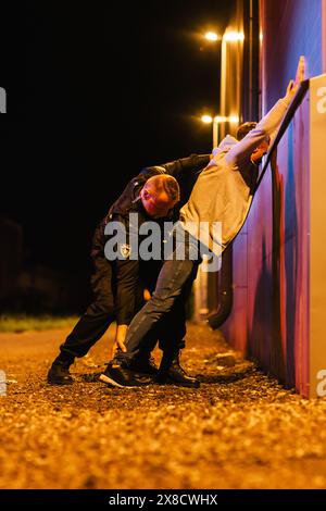 Un poliziotto professionista di mezza età che esegue una ricerca Pat-Down su un Fellon in Empty Back Alley. Un documentario come Shot of Procdure di arrestare i sospettati. Poliziotto esperto in cerca di armi Foto Stock