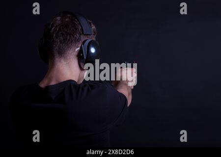 Giovane con protezione per orecchie e occhi che spara una pistola su sfondo nero Foto Stock