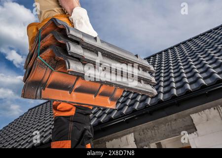 Il lavoratore caucasico del conciatoio edile è in piedi su un tetto, reggendo tegole in ceramica. Foto Stock