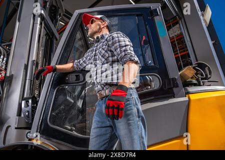 Un lavoratore in abbigliamento da lavoro si trova di fronte a un carrello elevatore in un ambiente industriale. Foto Stock