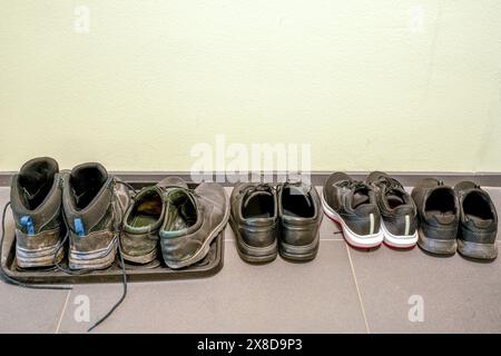 Scarpe, parzialmente usurate e sporche, in piedi di fila su un pavimento piastrellato, di fronte a una parete. Foto Stock