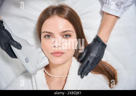 Vista dall'alto dell'estetista ritagliato che depilano il viso di una giovane donna al laser Foto Stock