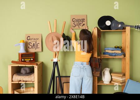 Giovane donna appesa cartone con VENDITA GARAGE testo a parete in camera di oggetti indesiderati, vista posteriore Foto Stock