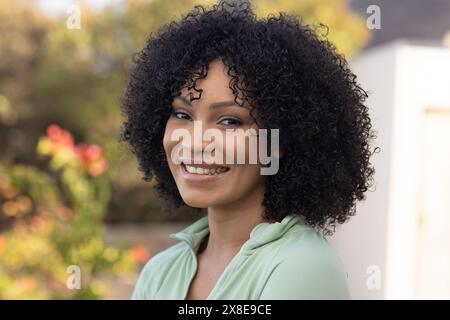 Una donna birazziale che indossa una blusa verde chiaro e si diverte all'aperto. Con i capelli neri ricci e la pelle marrone chiaro, sorride sotto un cielo blu limpido, unale Foto Stock