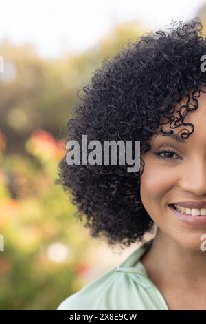 Una donna birazziale all'aperto, che indossa una camicetta verde chiaro, sorridente. Con i capelli neri ricci e la pelle marrone chiaro, che tiene il calore negli occhi scuri, godendosi Foto Stock