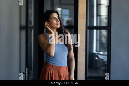 Nell'ufficio moderno, giovane donna indiana in piedi in casa, toccando l'orecchio. Ha lunghi capelli scuri, indossa un top senza maniche blu e pantaloni arancioni, con wa Foto Stock