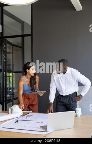 In un ufficio moderno, un team diversificato che lavora su piani architettonici e copia spazio. Giovane collega afroamericano che discute progetto con giovani indiani Foto Stock