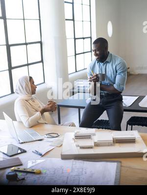 Team eterogenei in un ufficio moderno che discutono i dettagli del progetto e lo spazio di copia. Collega afroamericano che tiene in mano una tazza di caffè, mentre una donna indiana guida il team Foto Stock