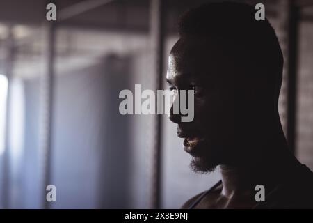 In palestra, forte e in forma uomo afroamericano sudato dopo un intenso allenamento, copia spazio. È in una palestra poco illuminata con attrezzature metalliche sullo sfondo, c Foto Stock