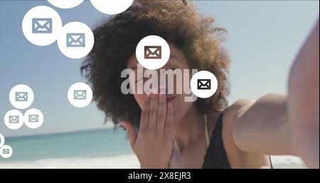 Immagine delle icone delle buste sopra una donna birazziale che tiene la macchina fotografica e bacio in spiaggia Foto Stock