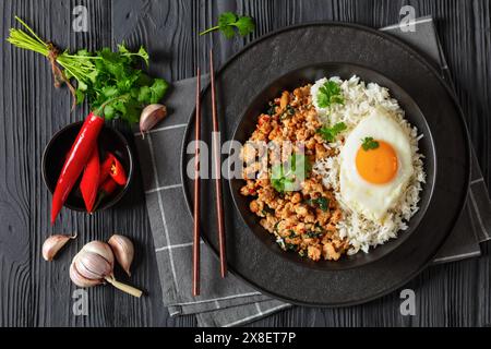 pad krapow gai, pad gaprao, pad kra pao, pollo al basilico fritto con riso e uovo fritto sul piatto con bacchette su un tavolo di legno nero, cucina tailandese, Foto Stock