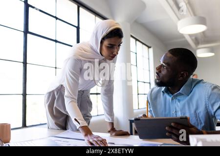 In un ufficio moderno, un team diversificato lavora insieme, discutendo disegni di architetti. Giovane afroamericano che tiene la tavoletta, giovane donna indiana in bianco Foto Stock