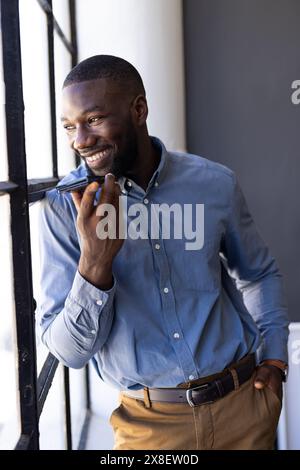 In un ufficio moderno, un giovane afroamericano in piedi davanti a una finestra, sorridente e parla. Capelli neri corti, barba rifinita, camicia con bottoni blu e marrone Foto Stock