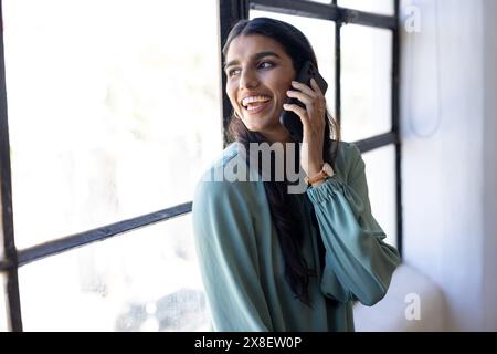 donna indiana con lunghi capelli neri e pelle leggera parla sullo smartphone in un ufficio moderno. Indossa una camicetta verde chiaro e guarda sorridendo mentre guardi fuori Foto Stock