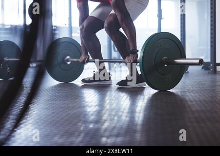 Uomo afroamericano forte e in forma che solleva il barbell in palestra. Circondato da attrezzature da palestra e pesi, concentrandosi sul suo allenamento con un look deciso, u Foto Stock