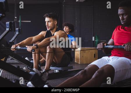 In palestra, tre giovani uomini diversi che si allenano con le macchine a remi. Sono in una moderna palestra con varie attrezzature fitness e sfondo scuro, inalterato Foto Stock