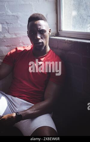 Uomo afroamericano forte e in forma, ventenne in palestra, con una camicia rossa. La luce del sole scorre attraverso una finestra, gettando ombre sullo sfondo di mattoni rossi Foto Stock