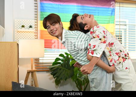 Ridendo la coppia gay maschio che si fa un giro in piggyback l'uno per l'altro in salotto con la bandiera arcobaleno dell'orgoglio LGBT Foto Stock