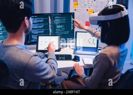 Lo sviluppatore di software asiatico che indossa un visore per la realtà virtuale lavora su un progetto di realtà virtuale, con un collega concentrato sul suo computer Foto Stock