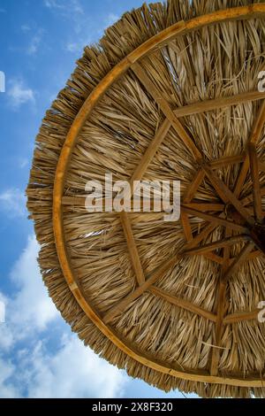 Una prospettiva unica che guarda da sotto un ombrellone di paglia, mostrando il suo intreccio intricato sullo sfondo di un cielo azzurro limpido con un morbido c Foto Stock