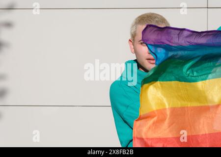 Un giovane uomo latino ha una bandiera arcobaleno ed è parzialmente coperto da essa. È un maschio biondo argentino, sta all'aperto con un muro sul dorso Foto Stock