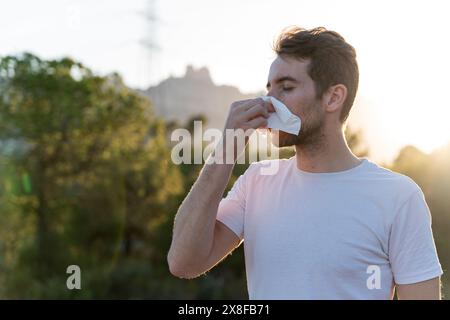 Uomo malato circondato dalla natura che soffia il naso e starnuto per i sintomi di allergia al polline Foto Stock