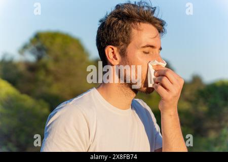 Uomo malato circondato dalla natura che soffia il naso e starnuto per i sintomi di allergia al polline Foto Stock