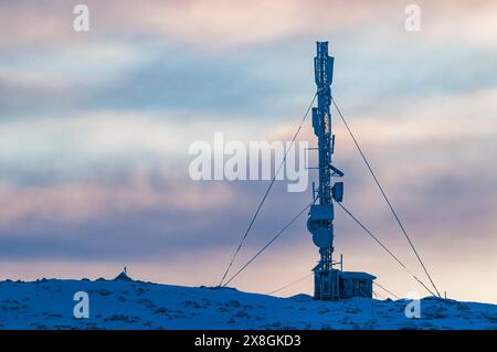 Una torre radio si erge in cima a una collina coperta di neve, in contrasto con il paesaggio bianco. La torre sembra robusta e industriale nel vento Foto Stock