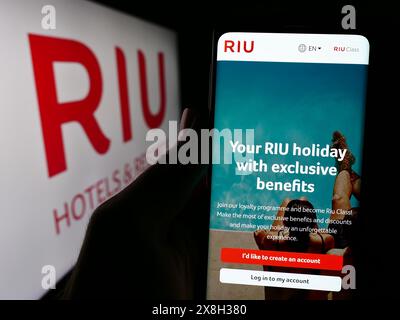 Persona che detiene un cellulare con pagina web della società alberghiera spagnola RIU Hotels and Resorts davanti al logo. Messa a fuoco al centro del display del telefono. Foto Stock