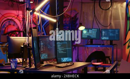 Linguaggio di programmazione sugli schermi dei PC in una base operativa vuota e disordinata di hacker con disegni grunge graffiti spruzzati sulle pareti. Nascondiglio al neon illuminato ghetto utilizzato dai criminali per commettere attività illegali Foto Stock