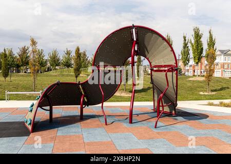 Moderne attrezzature da gioco - rosse e grigie - adagiate su piastrelle di gomma colorate - case suburbane e vegetazione sullo sfondo. Presa a Toronto, Canada. Foto Stock