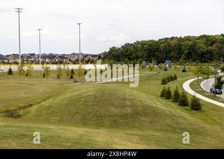 Parco suburbano con verdi colline ondulate - vialetti pavimentati e giovani alberi - parco giochi - sfondo case residenziali - pali luminosi alti e. Foto Stock